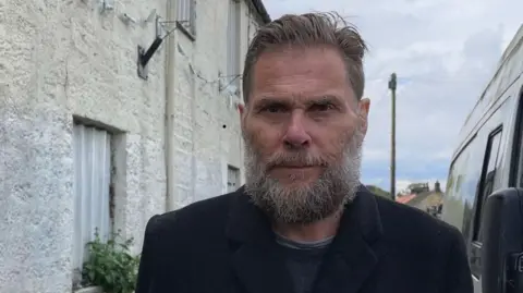 Man with a grey beard and moustache and short mousy hair wearing a black jacket and grey shirt is stood outside the pub 