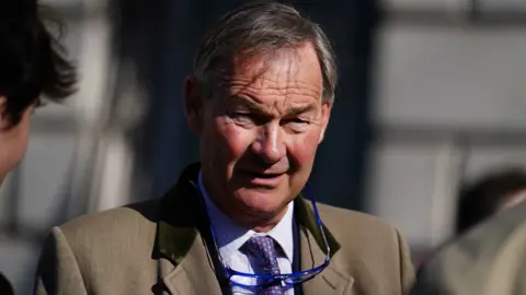 PA Media Rupert Lowe in a light brown blazer looks into the sun with a pair of glasses on a cord around his neck