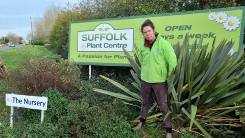 Contributed Harriet Lawton stands outside a garden centre entrance by a road. She stands in front of a green sign that reads Suffolk Plant Centre. She wears a green coloured jumper and burgundy trousers with brown boots.