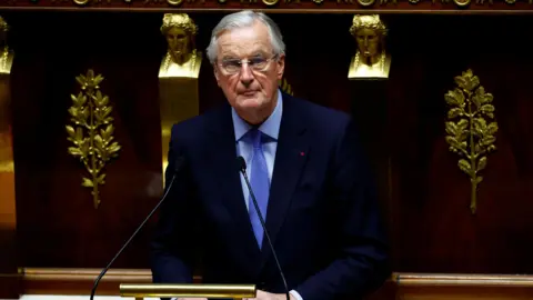 Reuters Michel Barnier, who has grey combed hair and wears spectacles with a navy suit and blue shirt, stands in front of mics at a lectern in the National Assembly in Paris