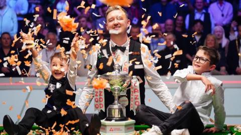 Kyren Wilson smiling behind a silver trophy while confetti falls from the roof