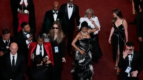 Actor and singer Teyana Taylor wearing a fitted gown of black and white feathers as she shimmies down the red carpet of the Acadamy Awards surrounded by other attendees. 