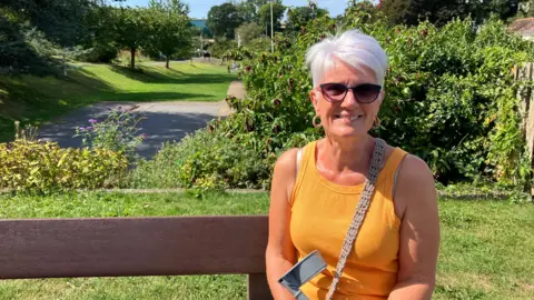 A woman with short white hair, wearing sunglasses and a yellow tank top. She is sat on a bench. The grass is green behind her. There are trees in the background. The sky is blue.