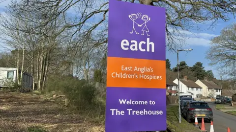 A large roadside sign for East Anglia's Children's Hospices, also known as EACH. The sign welcomes visitors to a facility called The Treehouse. It stands on a grassy verge beside a road.
