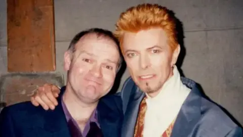 Angela Cambridge John stands on the left of the picture, David Bowie to the right. Bowie has his arm around John. He is smiling. Bowie has bright ginger hair, spiked up, while wearing a navy suit.