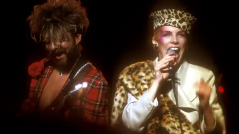 Getty Images Annie Lennox and Dave Stewart of the Eurythmics on stage in 1984. Dave wears a red tartan jacket and Annie is in a tailored camel trenchcoat with faux leopard fur wrap and matching hat.