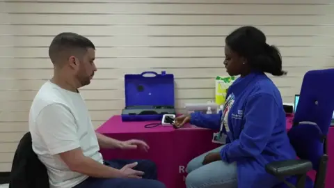BBC Wassim Chokri is sat opposite a woman at the new Community Wellness Outreach centre in Reading. The woman, who is wearing a blue fleece with the NHS logo, is preparing to do some tests on Chokri as part of a health check. She is holding a device and showing it to Chokri. 
