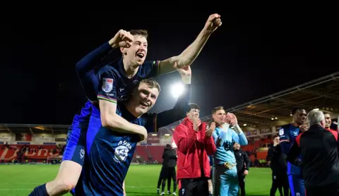 Chris Vaughan/Getty Images Two football players in dark blue kits celebrate on the pitch, with one player lifted on another’s back and raising an arm. Other players and staff stand nearby, clapping.