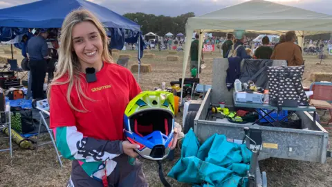 A woman, in her racing uniform and holding a helmet, is smiling at the camera. A dirt race track is behind her