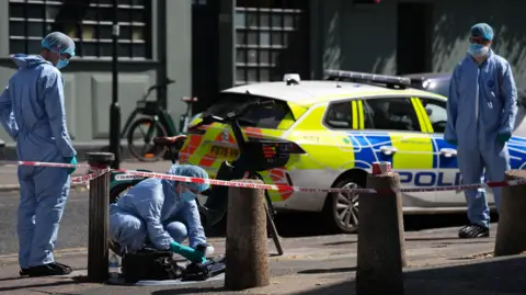 PA Forensic investigators in protective clothing examine a cordoned-off street scene near a bicycle, with police tape and a police car in the background.