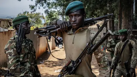 AFP A member of the M23 movement in military fatigues carries weapons over his shoulder. Another fighter is to his right and he is holding weapons under his arm.