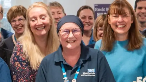 Kate Turner smiles at the camera surrounded by her colleagues. She wears a navy cover over her head, as well as a navy polo top with the West Suffolk NHS Foundation Trust logo on it. She also wears red glasses with a lanyard around her neck.