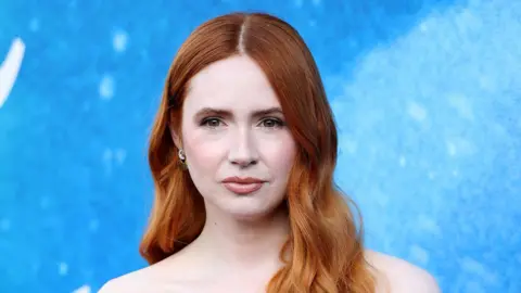 Getty Images Karen Gillan at "The Life of Chuck" Red Carpet Premiere held at the Hollywood Legion Theater. She has long wavy red hair and looks off to the side. There is a blue background behind her.