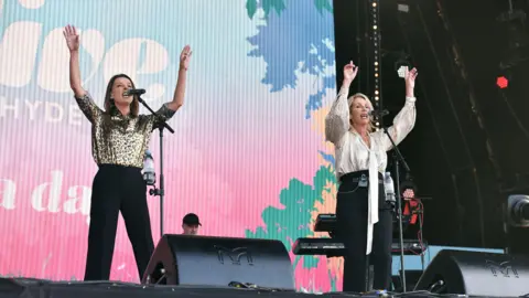 Bananarama - two women, one blonde and one brunette - hold their arms up in the air as they sing into microphones on a stage during a performance.