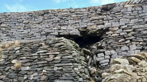 A wall which is made of hundreds of limestone bricks has collapsed. There is a large cave-sized hole that stems from the foundations. 