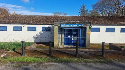 Stanhope Open Air Swimming Pool An exterior view of Stanhope Open Air Swimming Pool, a white single storey building with a late pitched roof and blue window frames. Gates and a fence made of blue metal bars prevent access to the central door. A sign above the door reads 'welcome to your pool'.