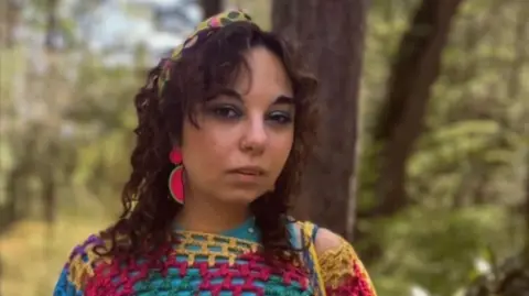 Saraya Haddad A woman wearing a multicoloured jumper and bright pink earrings with long brown hair stands in a forest.