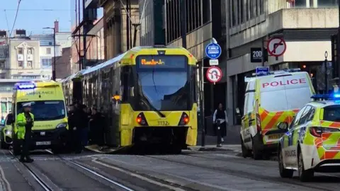 A stationary yellow and black Metrolink tram, flanked by an ambulance and police vehicles, at the scene of the collision.