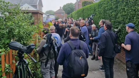 Andrea Blunt A crowd of men with cameras surround a residential garden