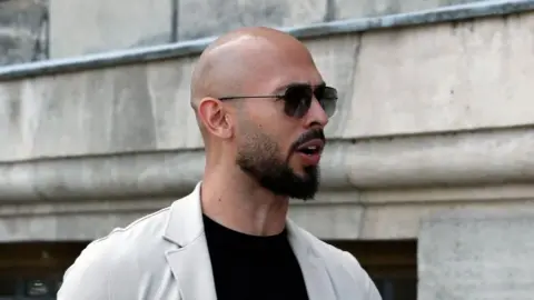 EPA-EFE/REX/Shutterstock Andrew Tate pictured outside, wearing a black T-shirt with a beige jacket and a pair of sunglasses.