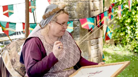 Mick Hurst A woman in a medieval costume and glasses is sewing. Behind her are bunting draped on a brick tower