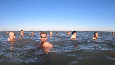 Mark Trigg Several men can be seen in a calm blue sea. One is looking to camera and has sunglasses on.