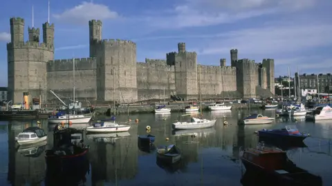 A huge stone castle wall jutting out onto the water surrounded by pleasure boats 