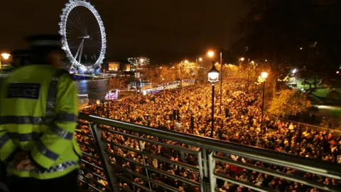 Getty Images Police officer in high-visibility jackets stand on a bridge overlooking a large crowd gathered along the Thames at night, with the London Eye lit up in the background.