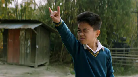 Excel Entertainment, Chalkboard Entertainment & Suitable Pictures A boy wearing a school uniform, doing a playful gesture with his fingers in an outdoor area.