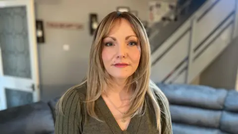 Abbie has long, blonde hair and a nose and lip piercing. She is looking directly into the camera and is wearing a green V-neck jumper and a silver necklace.