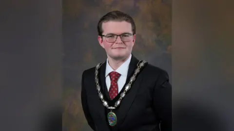 Alexander Walker photographed in a black three piece suit, white shirt and red tie. The 25-year-old has brown slicked back hair, square glasses, and a gold large livery chain. 