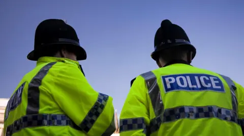 The back of two male police officers with blue sky in the background. The officers are wearing high-visibility green jackets with blue signs with white writing which says "police". 