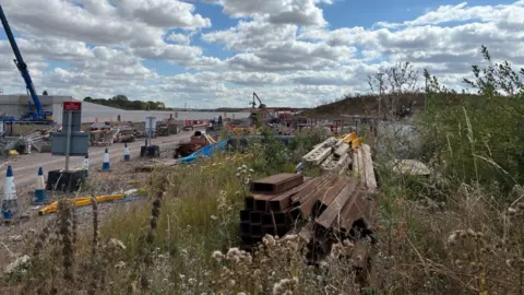 Una concurrida obra en construcción al aire libre, con grúas, maquinaria y materiales de construcción dispersos bajo un cielo nublado.