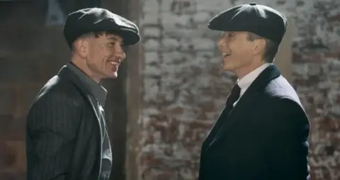 Netflix Duke Shelby, played by Saltburn star Barry Keoghan, stands opposite Tommy Shelby, played by Cillian Murphy. They are both wearing suits and flat caps and are clearly sharing a joke.