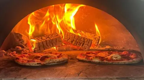 Copper Kiln Pizza Two circular and thin pizzas in a woodfired oven with fire blazing in the background