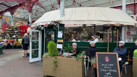 La Bouche restaurant in the Central Market in St Helier Jersey, a circular restaurant with a white roof, opposite a flower shop with an array of flowers in red, yellow, orange, white colours, people are sitting inside and outside of the restaurant with a sign in front which says the restaurant is open on Friday and Saturday nights