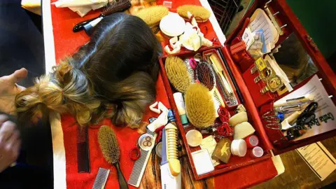 Getty Images A dog is sitting on a table covered with a red and white table cloth. The table is filled with brushes, combs and a red box filled with pens and hair accessories. 