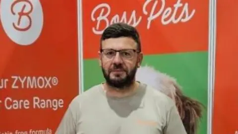 A man wearing glasses with a black beard stands in front of an orange banner which reads Boss Pets. 