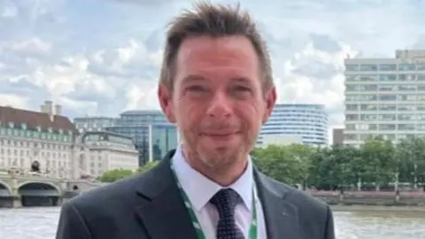 A man with short hair and wearing a dark suit, pale pink shirt, black tie and green lanyard around his neck, stands next to a river.