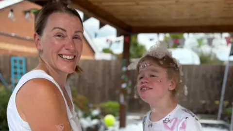 Family photo A woman with short brown hair and a white top and a small girl with blonde hair and a white tshirt in an outdoor area surrounded by fencing