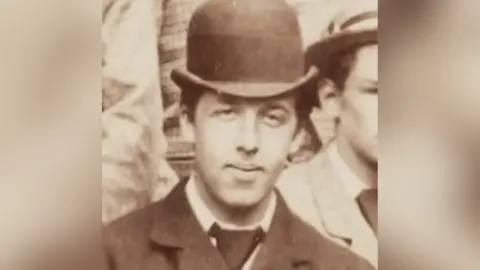 A black and white archive image of Oscar Wilde as a student. He is wearing a black bowler hat and suit.