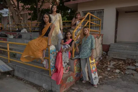 Chantal Pinzi Five young women stand on a concrete seating area which has yellow railings. four are holding skateboards