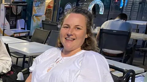 Sam Jennings Sam Jennings, who has long brown hair tied back from her face and is wearing a white top. She is looking into the camera and smiling. There are tables and chairs behind her