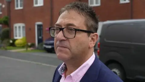 BBC Paul Carr wearing glasses and a pink shirt standing in a residential street