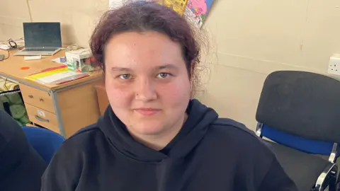A woman with purple dyed hair and a nose stud is sitting down in an office. She is wearing a dark hoodie.