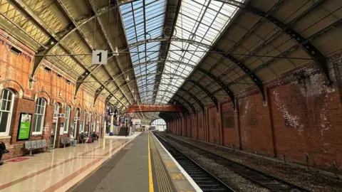 A view down one of the platforms of Darlington Station. The platfomr is wide and covered by a large arched metal work roof, supported by brick walls either side.