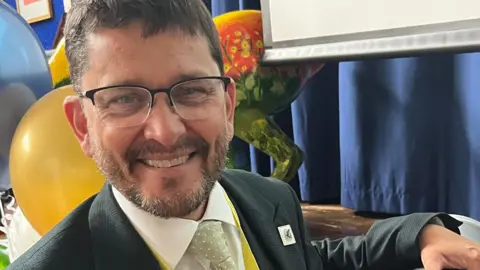 Craig Jensen has short brown hair and is wearing black framed glasses, a white shirt, black suit jacket and green tie. He is holding a silver metal time capsule to the right.