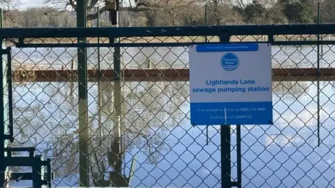 A metal fence with a Thames Water sign reading 'Lightlands Lane sewage pumping station' with a large reservoir of water behind it