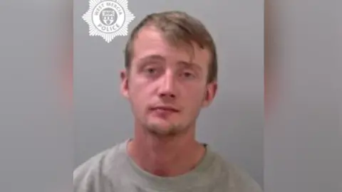 A man with brown hair and short facial hair is wearing a grey top in the photo. He is looing at the camera. There is a West Mercia Police logo in the top left corner of the image. 