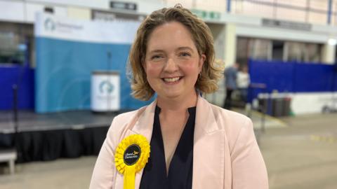 Anna Sabine smiling in a pink jacket, wearing a yellow lib dem rosette